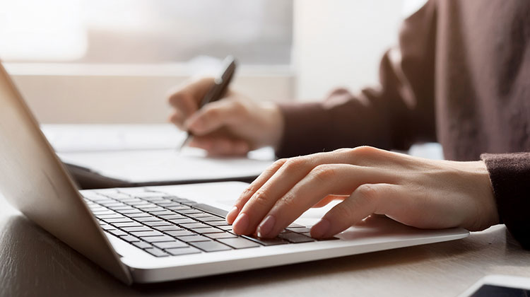 Man wearing brown top working at laptop and writing on paper