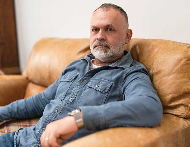 Man with grey hair in denim shirt sitting on a brown sofa