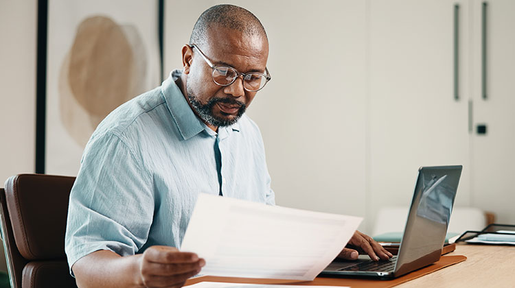 Man wearing blue shirt looking at sheet of paper while at his laptop