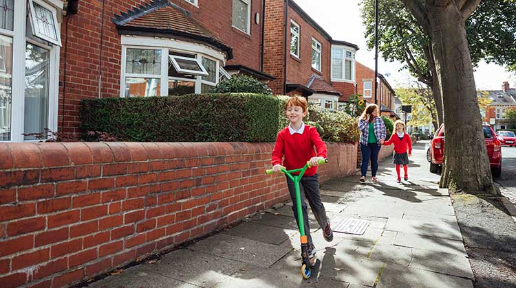 Child in red school jumper scootering on pavement with his mother behind him