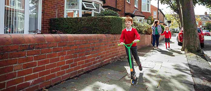 School boy in red jumper riding scooter with his family behind him