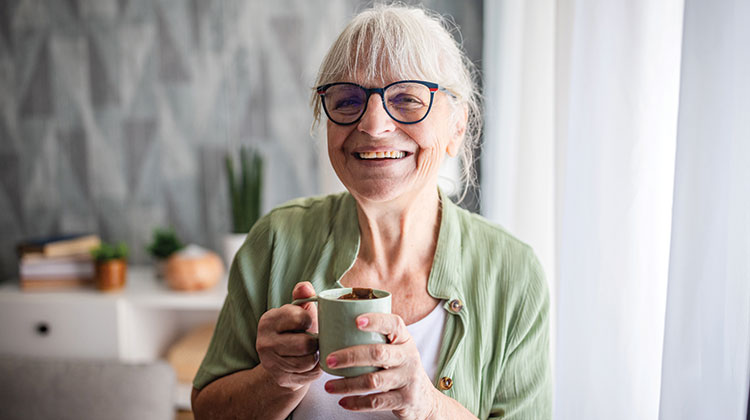 Smiling woman with grey hair in a ponytail, wearing glasses and a light green cardigan and holding a mug