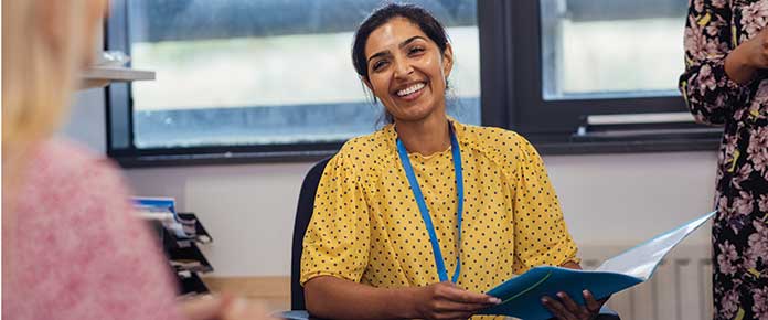 Woman wearing yellow top and lanyard card holder holding a file and talking to another woman