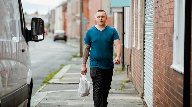 Man in teal t-shirt walking along street carrying a bag of shopping