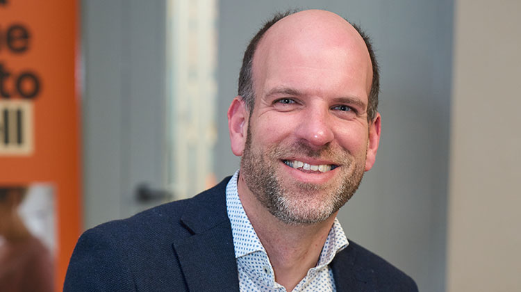 Sam Royston with short dark hair and beard wearing navy suit jacket and spotted shirt